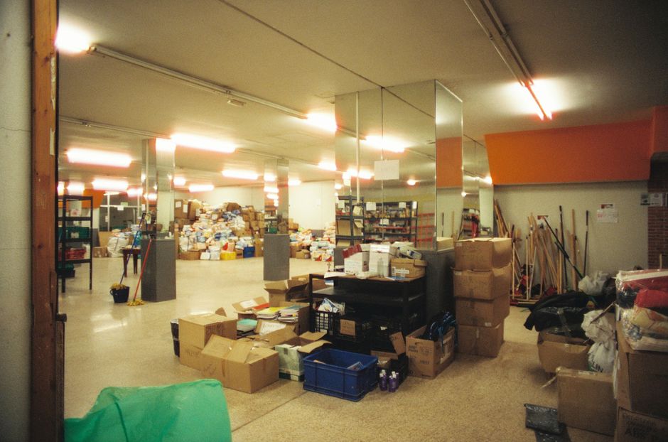 Large indoor storage area with stacks of boxes and supplies, illuminated by fluorescent lights.